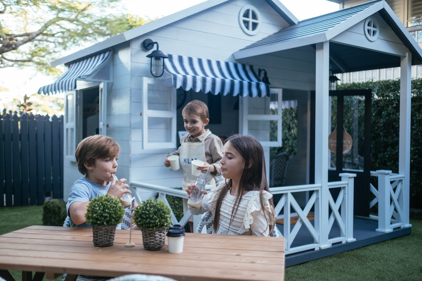 Children play cafe around a kids timber outdoor table. A white and black Cubby Cafe is in the background