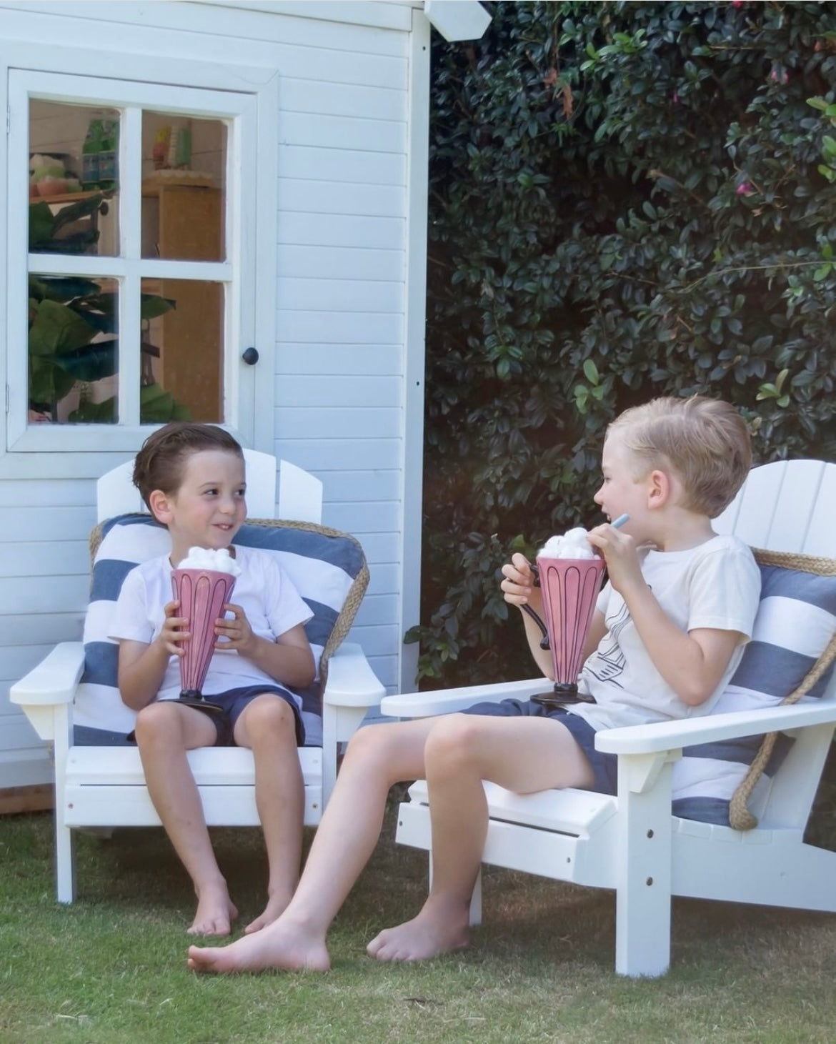 Two boys sitting outside in the garden sipping milkshakes on white children's Adirondack chairs