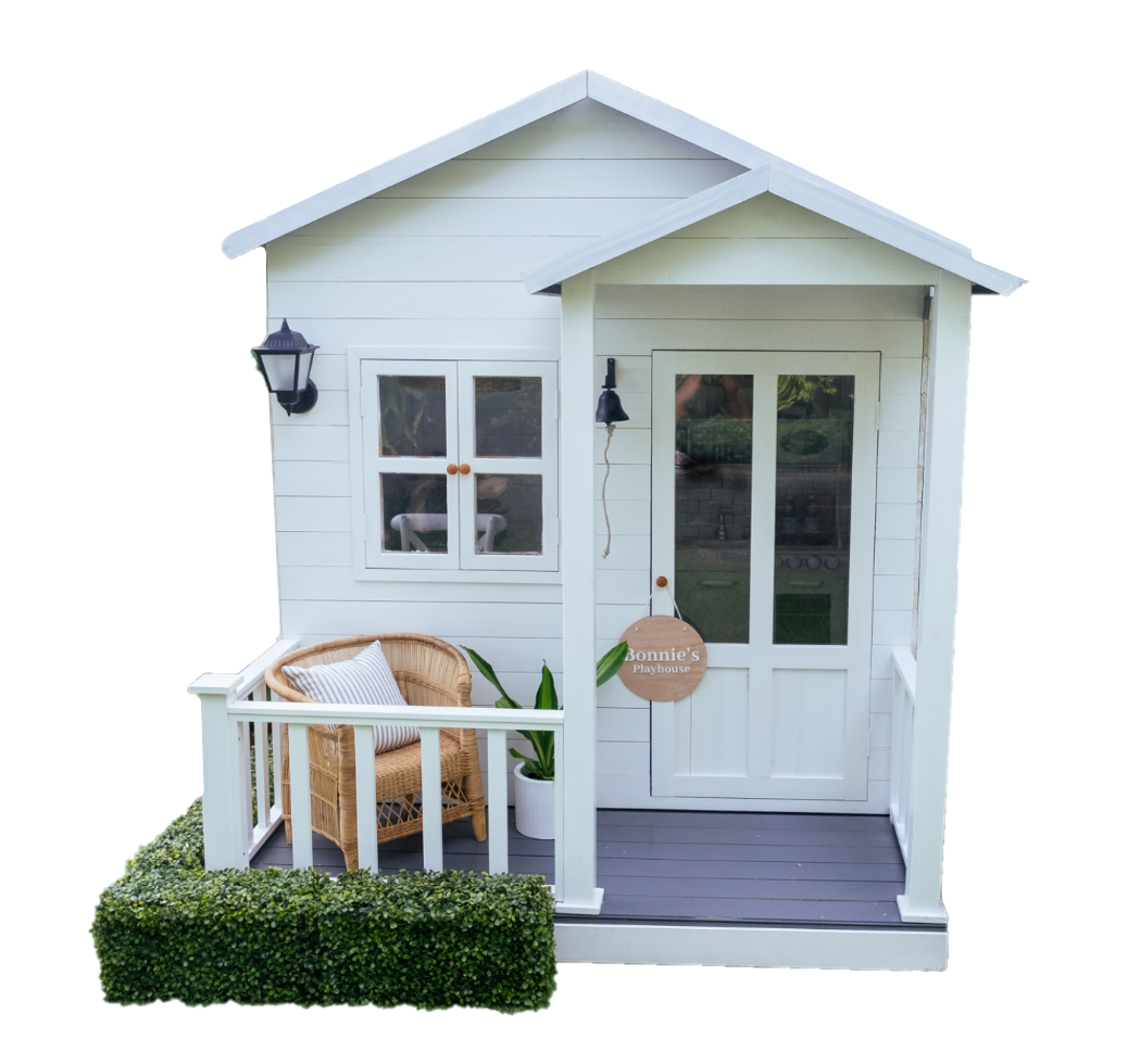 White cubby house with double gable roof and veranda sits on white transparent background. Kids Malawi arm chair sits on veranda