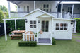 Large white hamptons cubby house has pergola and garage attachments. Kids outdoor furniture displayed