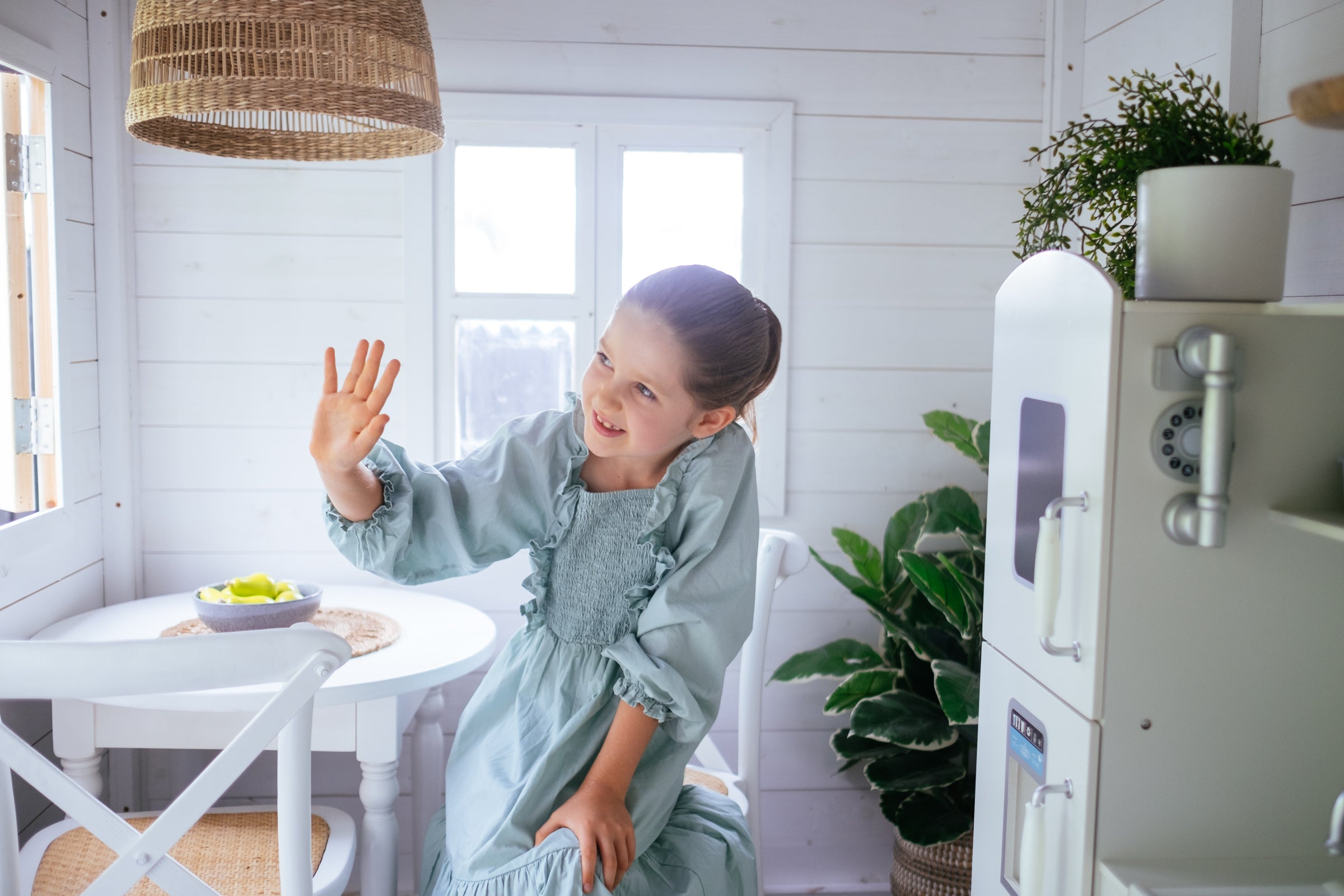 Young girl waves and smiles inside white timber cubby house. Kids white timber cross back chairs and white timber round table in corner.