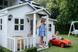 two young girls play in front of white timber cubby house. One stands on veranda the other is in small red car.