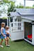 Two young girls walk towards white cubby house garage. A small red kids car sits inside.