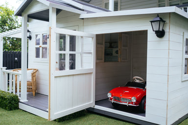 Small red car sits inside white timber cubby house garage.
