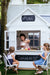 Two girls sit on kids bistro chairs in front of cubby house cafe. Cubby house is white and has blackboard signage.