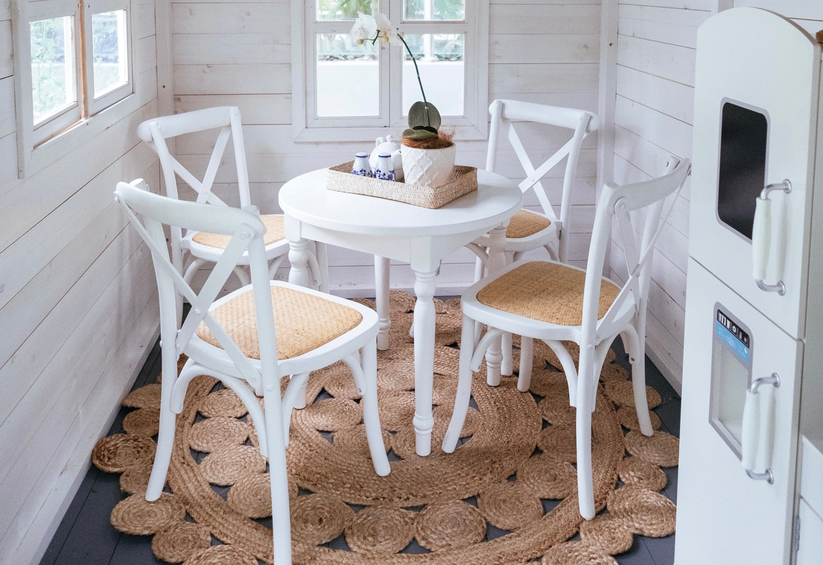 Kids Hamptons style cross back chairs in white and rattan sit around kids white round dining table inside timber cubby house