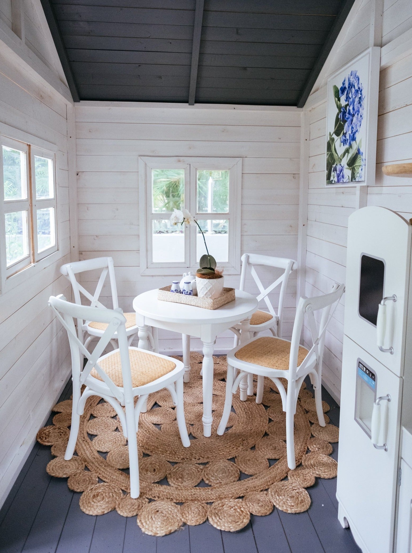 Kids cross back chairs and white round dining table sit on top of natural rug inside cubby house. The walls are white and the high ceiling is dark grey and matches floor. A floral print hangs from wall and the kids white play kitchen can be seen from the side