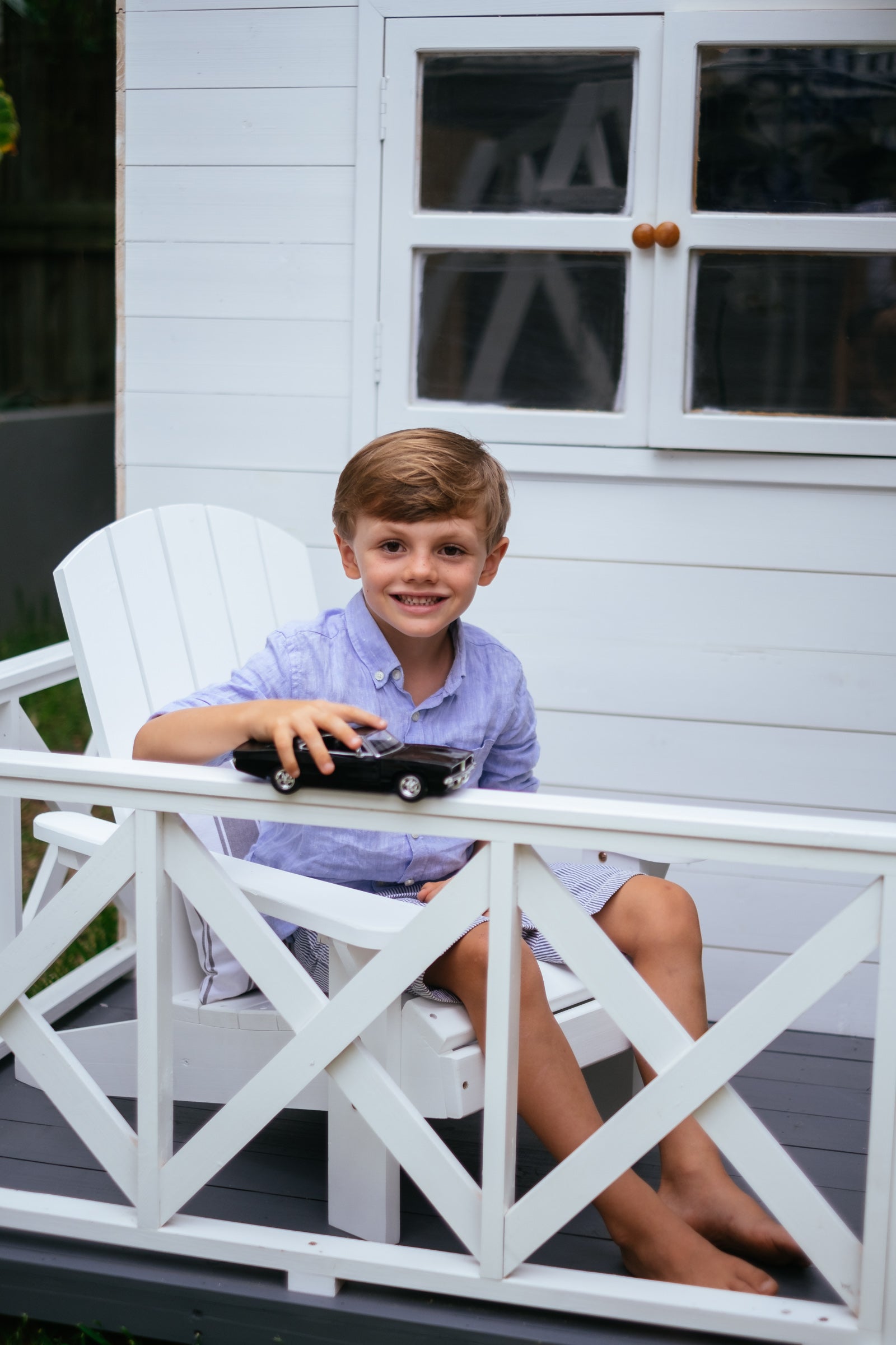 Young brunette boy sits on white timber kids Adirondack chair while playing with car.