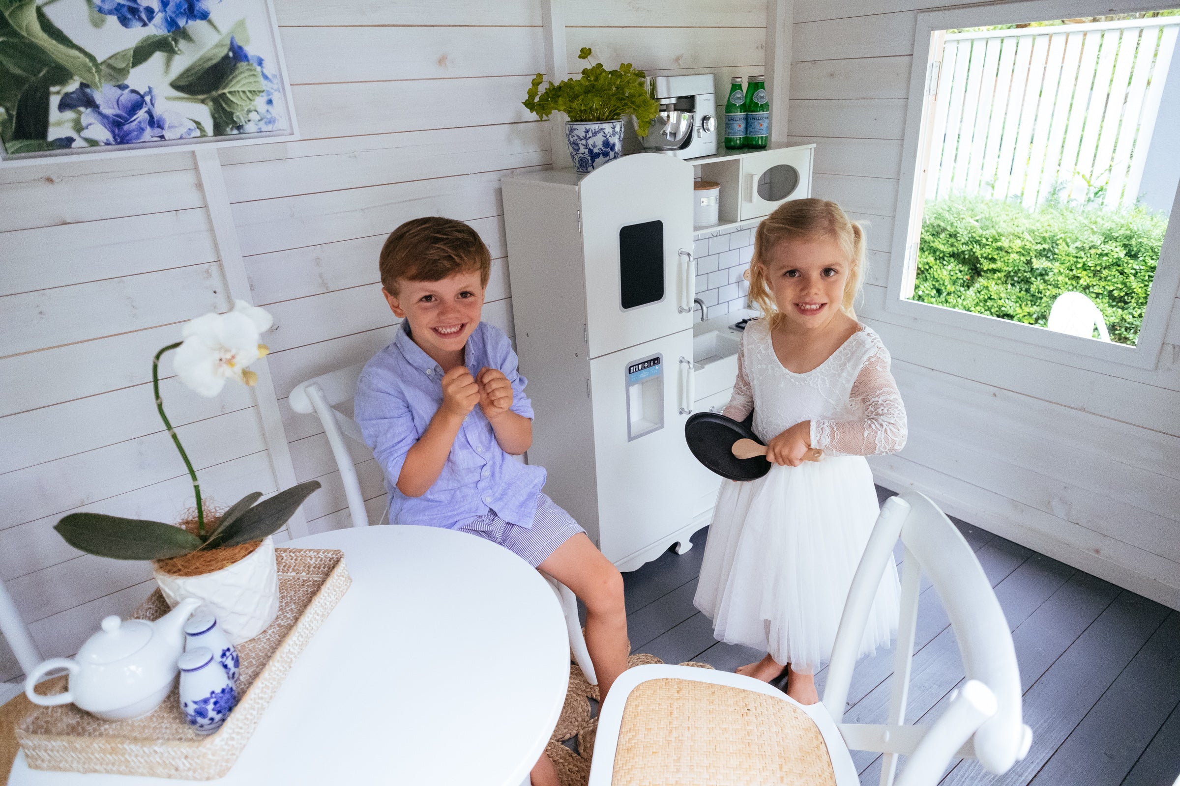 Young boy and girl play inside white and grey timber cubby house. White kids timber cross back chairs and white round dining table are in the corner