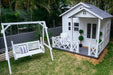 Large white Hamptons style cubby house next to white timber kids swing bench seat