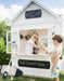 White cubby house with grey roof. Blackboard signage reads "Beach House. Surf Board Hire". A young blonde boy hangs out side window with yellow sunglasses and serves a pretend ice cream to young brunette boy wearing board shorts.